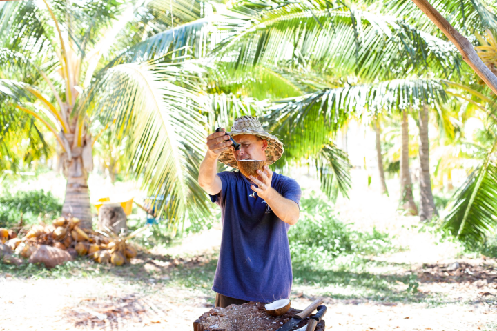 man in straw hat holding up half a coconut with a knife