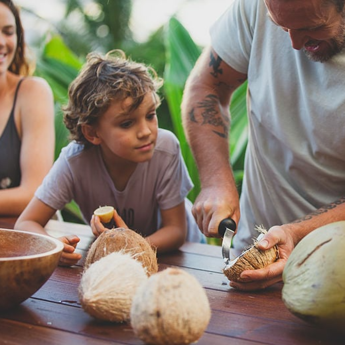 a woman and boy watching a man carve and harvest coconut meat