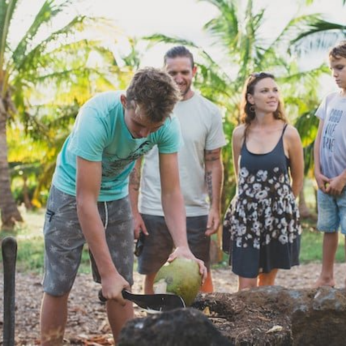 a group of people watching a teen cut the top of a coconut on a rock