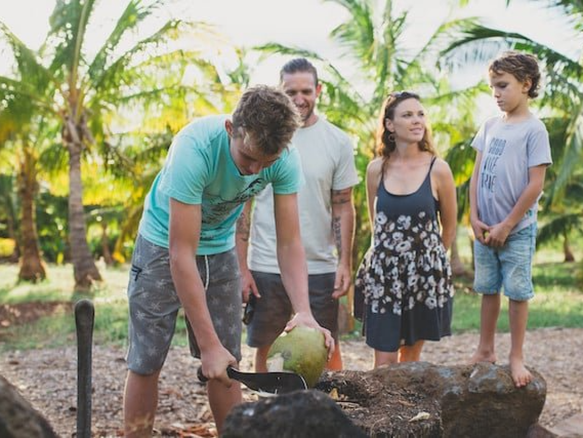 a group of people watching a teen cut the top of a coconut on a rock