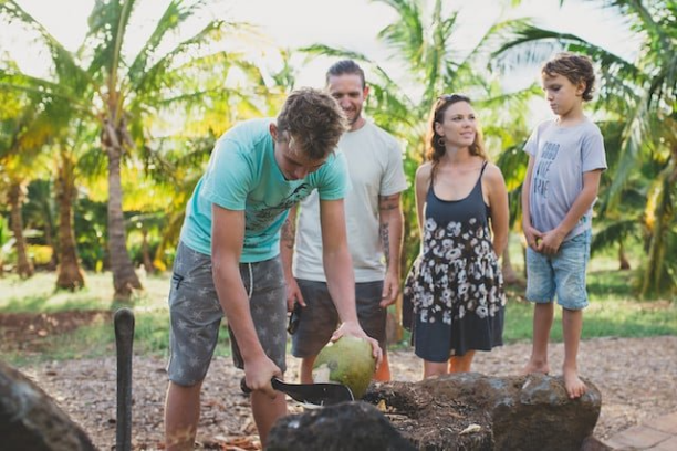 a group of people watching a teen cut the top of a coconut on a rock